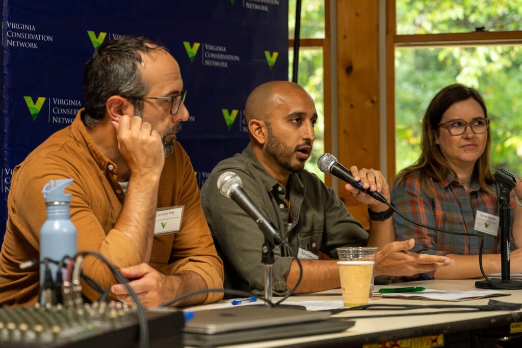 Three people sit on a panel with microphones in front of them. One person is talking.