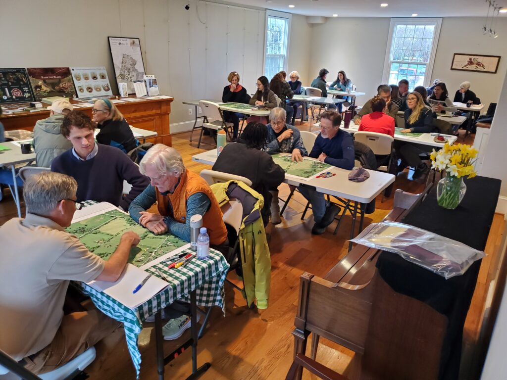 A room filled with people sitting across from each other at tables with maps in between them.