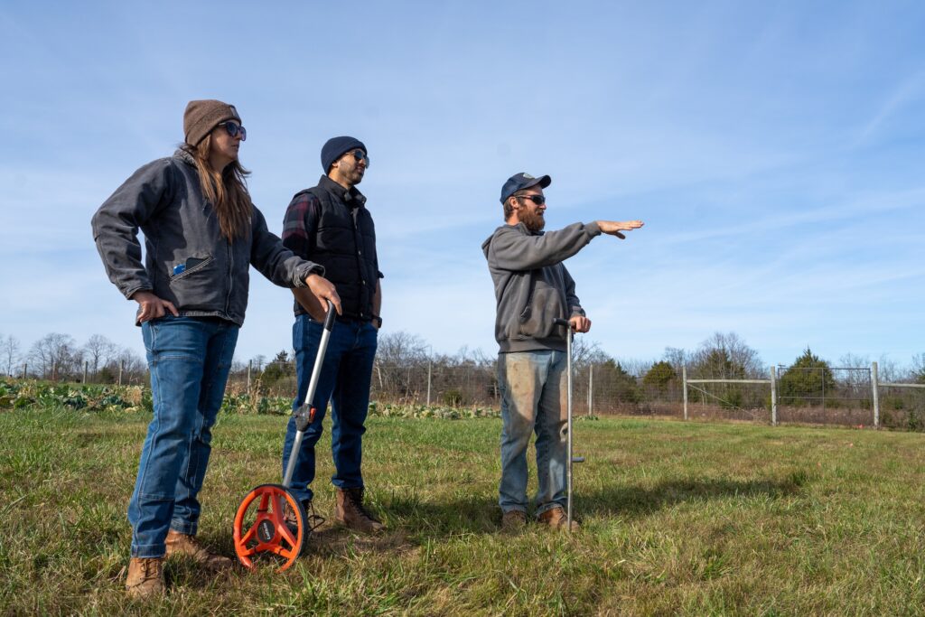 Three people stand in a field with surveying tools, looking out at the land in front of them. Crops are in rows behind them.